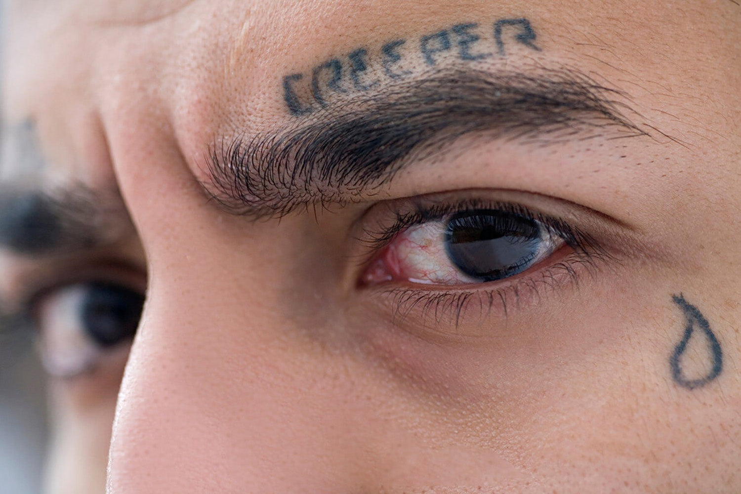 Acercamiento al rostro de una persona que tiene un tatuaje sobre la ceja de una palabra y una lágrima tatuada al lado de uno de sus ojos.