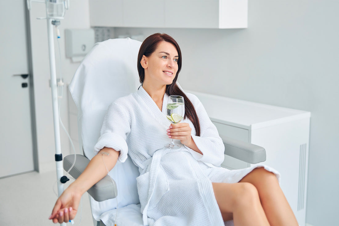 Mujer sonriendo y tomando agua de una copa mientras recibe un tratamiento cosmético por medio intravenoso.