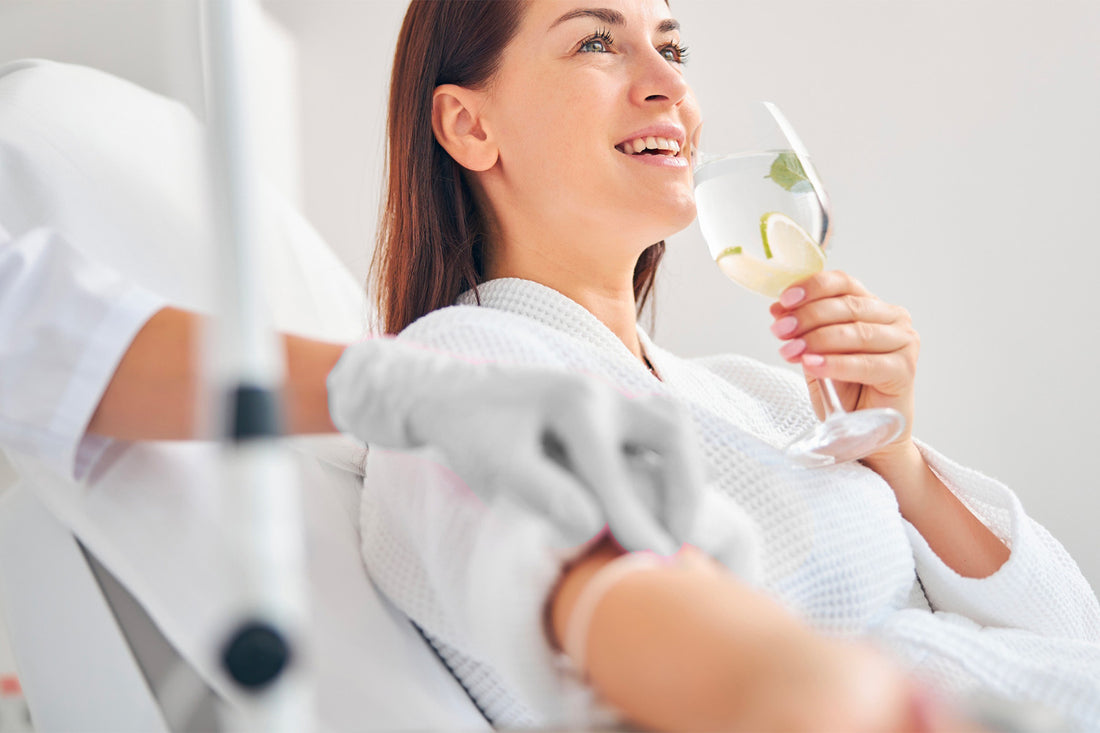 Mujer sonriendo y tomando agua de una copa mientras recibe un tratamiento cosmético por medio intravenoso.